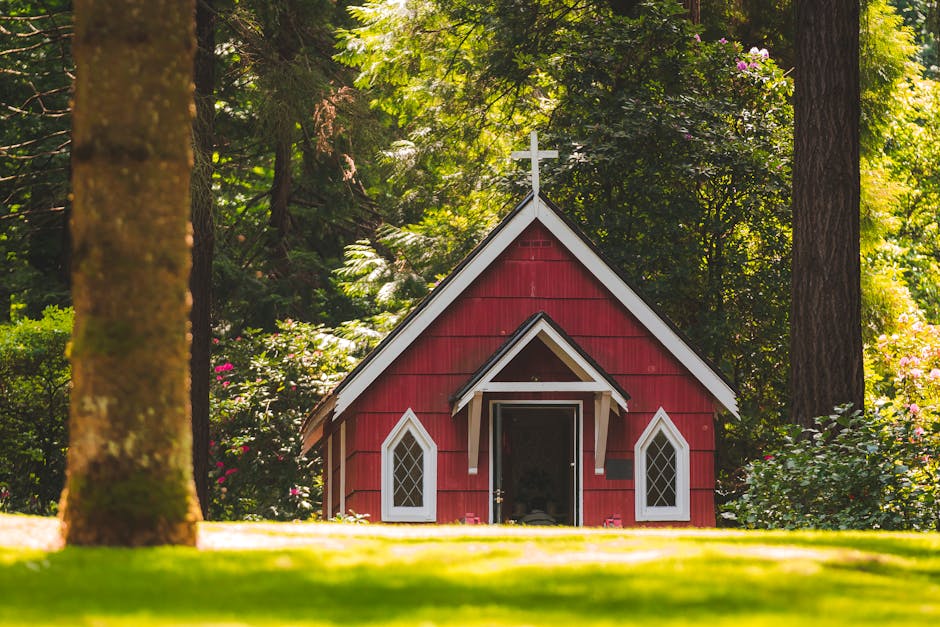 A small, red-painted wooden chapel with white trim and diamond-shaped pane windows, situated in a lush garden setting. The building features a steep gabled roof topped with a white cross and a central