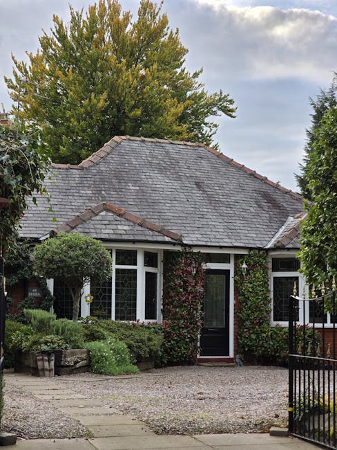 A small, red-painted wooden chapel with white trim and diamond-shaped pane windows, situated in a lush garden setting. The building features a steep gabled roof topped with a white cross and a central